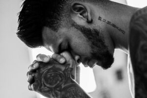 A man in deep thought and prayer, showcasing tattoos and an earring in a powerful black and white image.